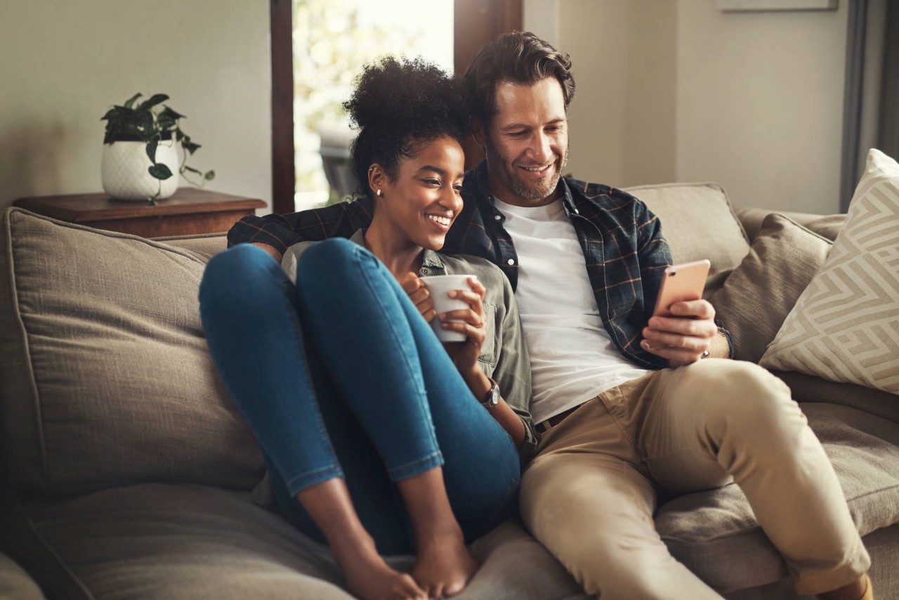 A couple sitting on their couch controlling their automatic blinds for windows from their phones at at Geneva’s Interiors near Federal Way, WA